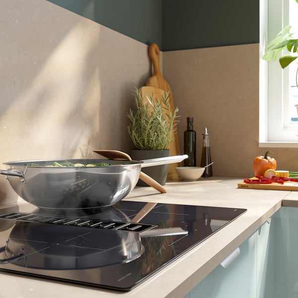 Moderne Küche mit Induktionskochfeld aus Edelstahl, Kochen von grünem Gemüse, Holzutensilien, Flaschen und frischen Produkten.