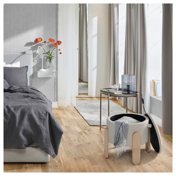 Modernes Schlafzimmer mit dunkler Bettwäsche, Holzböden, grauen Wänden und großen Fenstern. FÖRLUNDA Pouf in Weiß neben einem Tisch mit Blumen und Vasen.