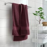 Badezimmer mit hängendem kastanienbraunem FREDRIKSJÖN-Handtuch, weicher und dicker Baumwolle, weiß gefliester Wand, kleiner grüner Pflanze auf dem Regal.