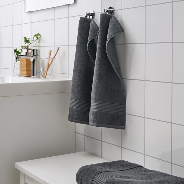 Dunkelgraue Fredriksjøn-Handtücher hängen und falten sich ordentlich an weiß gefliesten Badezimmerwänden und kontrastieren mit kleineren Gegenständen auf der Theke.
