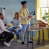 Drei Personen in einem gemütlichen blauen Zimmer. Einer hält eine Schallplatte, ein anderer Chips. Zwei Holztische mit Schallplatten und Essen. Eine Lampe erhellt die Szenerie.