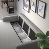Graues gepolstertes Schlafsofa mit offenem Staufach unter dem Sitz, auf dem Bettwäsche und Kissen ausgestellt sind. Umgeben von Wandkunst und Regalen mit Büchern.