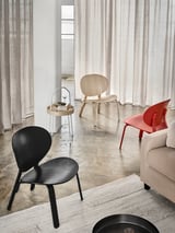 Modernes Wohnzimmer mit drei verschiedenen Stühlen: Schwarz, Naturholz und Rot, angeordnet um einen Teppich. Leichte Vorhänge und minimalistisches Dekor.