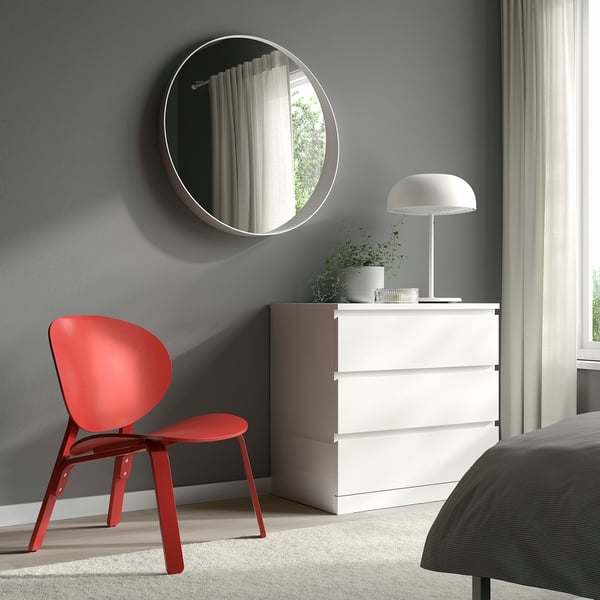 Modernes Schlafzimmer mit rotem FRÖSET-Stuhl, weißer Kommode, Pflanze und rundem Spiegel. Hellgraue Wände. Bett teilweise sichtbar. Effiziente Raumnutzung.