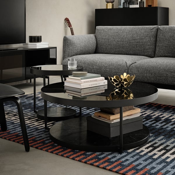 Modernes Wohnzimmer mit grauem Sofa, schwarzem Beistelltisch und buntem Teppich. Auf dem Tisch stehen Bücher, Glas und dekorative Kunst.