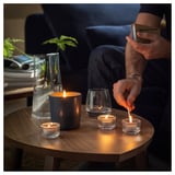 Die Person zündet eine Kerze neben einer brennenden Glaskerze, einem Weinglas und einer Vase mit Pflanze auf einem dunklen Holztisch an.