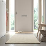 Beigefarbener Teppich an der Tür, Stuhl mit Kissen, Jacke an der Wand, heller Raum.