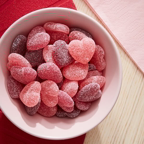 Weiße Schale mit rosafarbenen, mit Zucker überzogenen Herzbonbons. Bonbons sind rot und rosa und in der Schüssel verstreut. Schüssel auf einer Holzunterlage mit rotem Tuch.