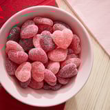 Weiße Schale mit rosafarbenen, mit Zucker überzogenen Herzbonbons. Bonbons sind rot und rosa und in der Schüssel verstreut. Schüssel auf einer Holzunterlage mit rotem Tuch.