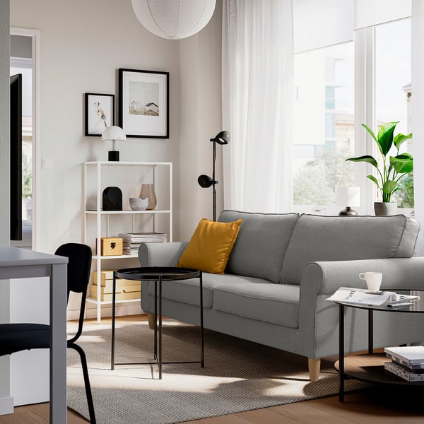 Modernes Wohnzimmer mit grauem GÄLLTOFTA-Sofa , gelbem Kissen, schwarzem Beistelltisch, weißen Vorhängen, großem Fenster und Pflanzen.