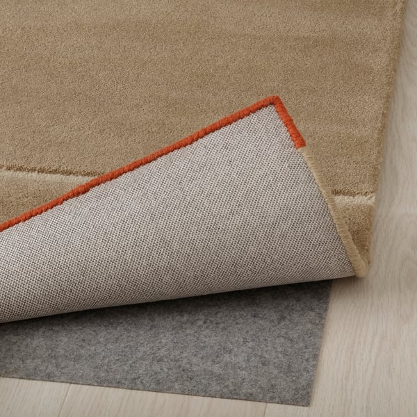 Teppich mit Wollstruktur, roter Rand, zurückgefaltet, um eine gefilzte Unterlage zu zeigen. Dieses Setup verhindert ein Verrutschen und erhöht den Komfort.