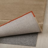 Teppich mit Wollstruktur, roter Rand, zurückgefaltet, um eine gefilzte Unterlage zu zeigen. Dieses Setup verhindert ein Verrutschen und erhöht den Komfort.