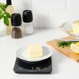 Schwarze digitale Küchenwaage mit weißem Teller und Butter; LED-Display, gehackte Kräuter, Zitrone und Messer in der Nähe.