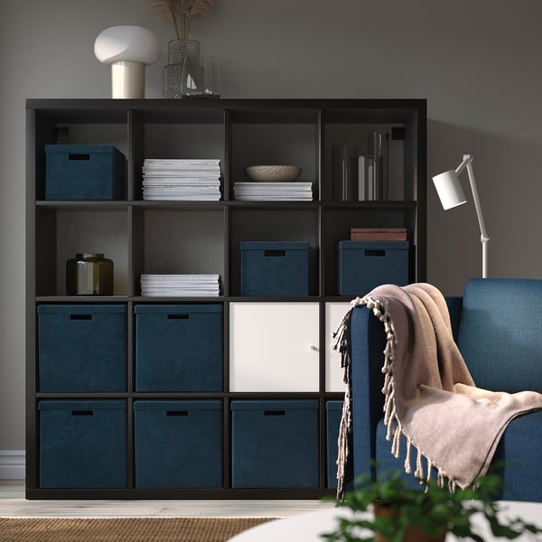 Modernes Wohnzimmer mit GJÄTTA-Aufbewahrungsboxen auf dunklem Regal, die mit Büchern und einem gemütlichen Stuhl verschmelzen.