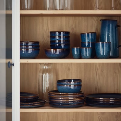 Holzregal mit blauen GLADELIG Bechern, Schalen, Tellern und Krug. Goldbraune Details, Steinzeug.