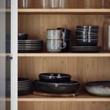 GLADELIG, graues Geschirr, Steinzeug, auf Regalen gestapelt. Größere Teller, Tassen, Schalen, einzigartiges Design.