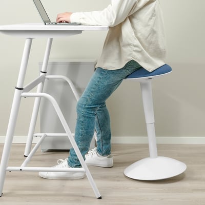 Person mit weißem Sitz-/Steh-Schreibtisch, tippt auf Laptop, während sie auf einem hohen Hocker mit blauem Sitz sitzt.