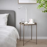 GLADOM schwarzer Beistelltisch mit weißer Lampe, Glas, Uhr, neben ordentlich gemachtem Bett mit weißer Bettwäsche, grauem Kopfteil und Holzboden.