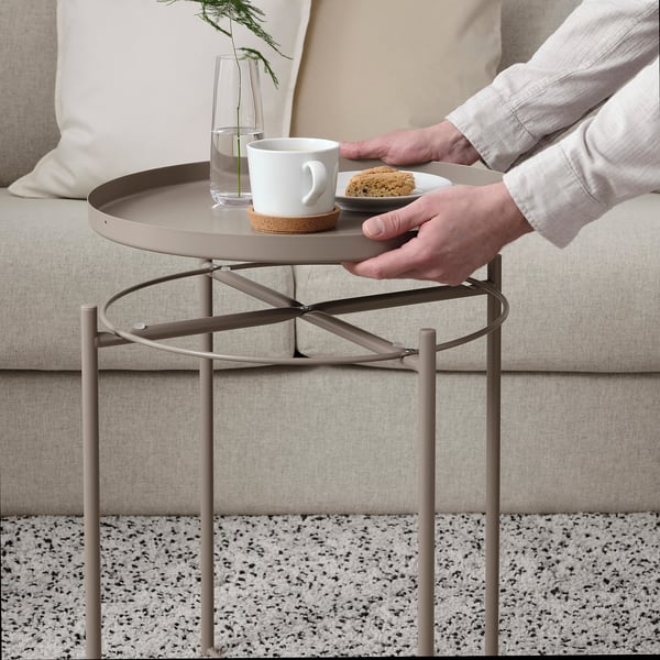 Person mit einer Kaffeetasse und einem Keks auf einem runden Metalltabletttisch neben einer Couch.