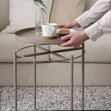 Person mit einer Kaffeetasse und einem Keks auf einem runden Metalltabletttisch neben einer Couch.