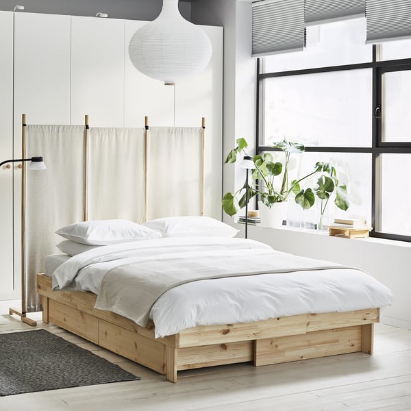 Modernes weißes Schlafzimmer mit ausziehbarem Bett aus hellem Holz, durchsichtigen Vorhängen, großen Fenstern und grüner Pflanze.