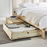 Modernes Schlafzimmer mit ausziehbarem Bett aus hellem Holz GLAMBERGET, weißer Bettwäsche und offenen Schubladen, in denen gefaltete Kleidung zu sehen ist.