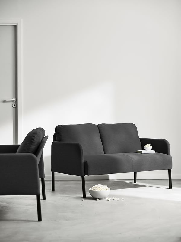 Das schwarze, moderne Sofa mit Armlehnen und Rückenkissen lehnt sich an eine schlichte Wand. Eine kleine Schüssel und verschüttetes Popcorn auf dem Boden daneben.