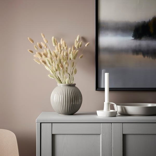 Ein minimalistischer, moderner Kerzenständer namens GODTAGBAR, weiß in der Farbe, aus Steinzeug, klein und schlank neben einer großen Vase auf grauer Oberfläche.