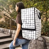 Eine Person, die einen GÖRSNYGG Rucksack in Schwarz und Weiß trägt, der größer als typische Rucksäcke ist und für das Tragen oder Bewegen großer Lasten konzipiert ist.