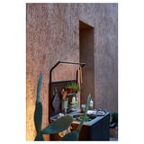 Außenkücheneinrichtung mit Edelstahlspüle und Regalen aus verzinktem Stahl an strukturierter Wand. Verschiedene Küchenhelfer und Pflanzen ordentlich angeordnet.
