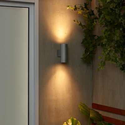 Moderne graue Außenwandleuchte GRÖNSPRÖT an beiger Wand in der Nähe der weißen Tür, mit warmem Licht und Grünkontrast.
