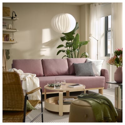 Modernes Wohnzimmer mit grünem, lavendelfarbenem Schlafsofa, hölzernem Couchtisch, Regalen und großer Pflanze unter einer weißen Pendelleuchte.