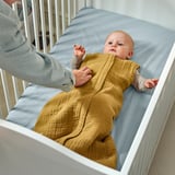 Ein Baby in einem gelben Schlafsack liegt in einem Bettchen, die Hand eines Erwachsenen in der Nähe.