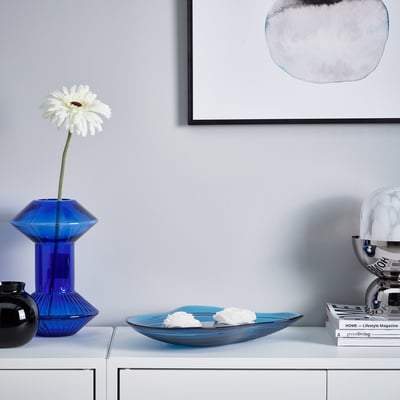 Moderne Tischaufstellung: blaue Schale, hohe Vase mit weißer Blume, schwarze Schale, Zeitschriften, graue Wand, runde Kunst.