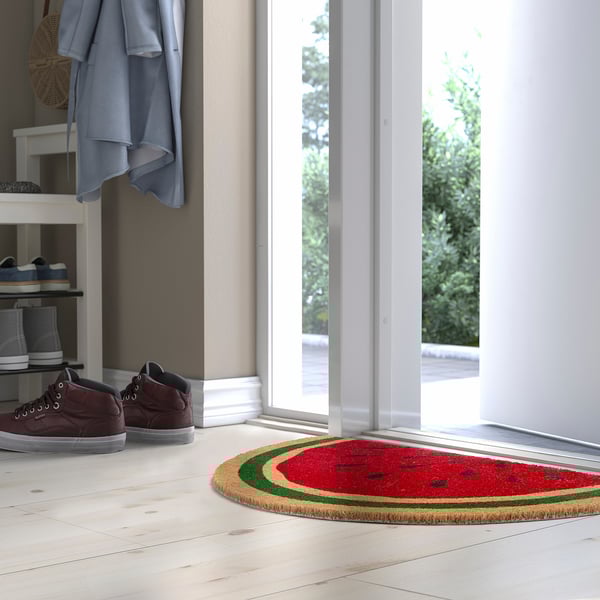 Leuchtend rote wassermelonenförmige Fußmatte mit grüner Rinde unter einem Schuhregal, neben einem blauen Mantel und einer Tür.