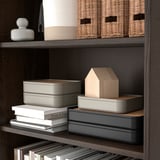 Organisiertes Regal mit grauen Kisten, Weidenkörben, Büchern und Holzdekor.