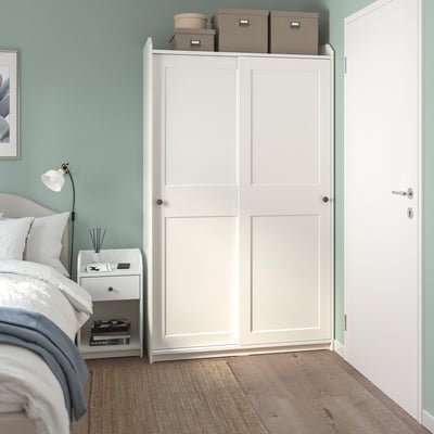 Modernes Schlafzimmer mit hohem weißem HAUGA-Kleiderschrank, Bett, Nachttisch, Lampe und Büchern. Hellblaue Wände, beruhigende Atmosphäre.
