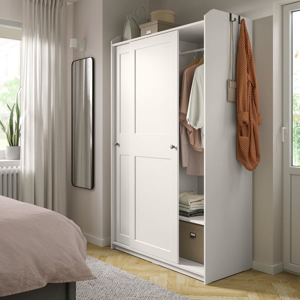 Ein weißer Kleiderschrank von HAUGA mit Schiebetüren in einem Schlafzimmer. Eine Tür ist offen und gibt den Blick auf Kleidung und Aufbewahrungsboxen frei. Ein Spiegel und eine Pflanze befinden sich in der Nähe.