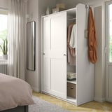 Ein weißer Kleiderschrank von HAUGA mit Schiebetüren in einem Schlafzimmer. Eine Tür ist offen und gibt den Blick auf Kleidung und Aufbewahrungsboxen frei. Ein Spiegel und eine Pflanze befinden sich in der Nähe.