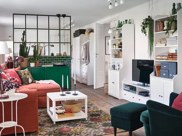 Helles Wohnzimmer mit weißem Stauraum von HAUGA, grünen Akzenten, rotem Sofa, üppigen Pflanzen und buntem Teppich. TV-Ständer, Regale mit Büchern und Dekoration.