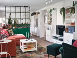 Helles Wohnzimmer mit weißem Stauraum von HAUGA, grünen Akzenten, rotem Sofa, üppigen Pflanzen und buntem Teppich. TV-Ständer, Regale mit Büchern und Dekoration.