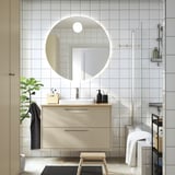 Modernes Badezimmer mit großem Spiegel, beigem Schrank, halb versenktem Waschbecken, Holzhocker, schwarzem Handtuch, Glasdusche, Pflanze.