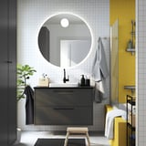Modernes Badezimmer mit rundem Spiegel, grauem schwebendem Waschtisch, weißem Waschbecken, gelben Akzentfliesen, Holzhocker, Pflanze, Handtüchern und Glasdusche.