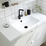 Modernes weißes Badezimmer mit HAVBÄCK-Waschtisch, zwei Schubladen, elegantem Waschbecken, schwarzem Wasserhahn, Seifenspender, Pflanzen- und Zahnbürstenhalter.