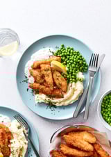 Ein Teller mit knusprig goldenem Fisch beißt auf cremigem Brei mit grünen Erbsen und Zitrone. Zwei Gabeln und ein Glas Limonade an der Seite.
