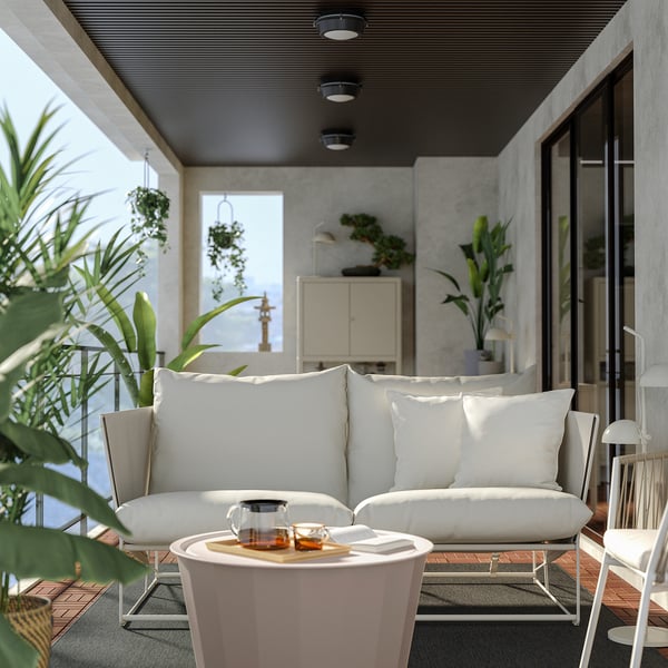 Modernes Outdoor-Sofa mit weißen Kissen, Metallrahmen und Glastisch, umgeben von Pflanzen auf einem gemütlichen Balkon.