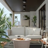 Modernes Outdoor-Sofa mit weißen Kissen, Metallrahmen und Glastisch, umgeben von Pflanzen auf einem gemütlichen Balkon.
