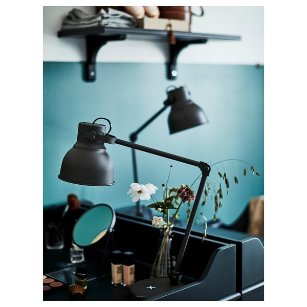Modernes Schlafzimmer mit schwarzen, verstellbaren HEKTAR-Lampen, eine auf einem Wandregal und eine auf einem Schreibtisch neben einem blauen Spiegel und einer Vase mit Blumen.