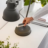 Die Person lädt das Smartphone über ein kabelloses Ladegerät auf dem Schreibtisch mit verstellbarer Lampe auf.