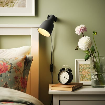 Die HEKTAR Wandleuchte beleuchtet den Nachttisch mit Blumen und Uhr, neben dem Bett mit Kissen.